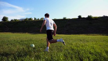 Üniformalı profesyonel futbolcu yeşil sahada koşarken topa numara yapıyor. Sporcu güneşli bir günde Meadow 'da koşarken topa vurur. Stadyumda antrenman yapan genç adam. Serbest stil futbol.