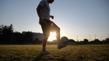 Günbatımında stadyumda futbol topu hokkabazlığı yapan profesyonel bir futbolcu. Genç adam yeşil sahada topa vuruyor. Sporcu arka planda güneş ışığıyla çayırda numaralar yapıyor. Serbest stil futbol kavramı.