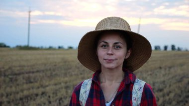 Mutlu gülümseyen dişi çiftçi buğday tarlasında kameraya bakıyor. Hasır şapkalı, arka planda arpa çayırı olan genç ve güzel tarımcı portresi. Tarım sektörü kavramı.
