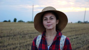 Mutlu gülümseyen dişi çiftçi buğday tarlasında kameraya bakıyor. Hasır şapkalı, arka planda arpa çayırı olan genç ve güzel tarımcı portresi. Tarım sektörü kavramı.