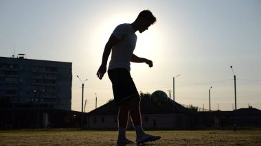 Günbatımında stadyumda futbol topu hokkabazlığı yapan profesyonel bir futbolcu. Genç adam yeşil sahada topa vuruyor. Sporcu arka planda güneş ışığıyla çayırda numaralar yapıyor. Serbest stil futbol kavramı.