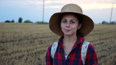 Mutlu gülümseyen dişi çiftçi buğday tarlasında kameraya bakıyor. Hasır şapkalı, arka planda arpa çayırı olan genç ve güzel tarımcı portresi. Tarım sektörü kavramı.