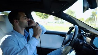 Şehir yolunda otomatik pilotla elektrikli otomobil sürerken akıllı telefondan konuşan erkek girişimci. Kendine güvenen işadamı, otomatik elektrikli araba kullanırken telefonla konuşuyor..