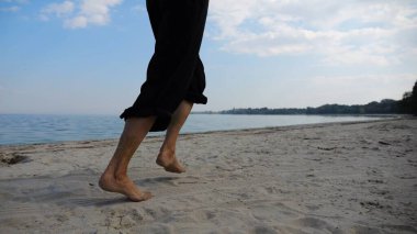 Sahilde arka planda deniz manzarasıyla koşan çıplak ayaklı bir kadın. Deniz kıyısında koşan dişi bacaklar. Mutlu bayan okyanus kıyısında vakit geçiriyor. Yaz tatili kavramı.