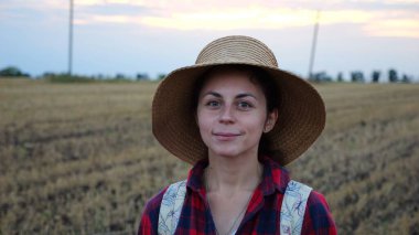 Mutlu gülümseyen dişi çiftçi buğday tarlasında kameraya bakıyor. Hasır şapkalı, arka planda arpa çayırı olan genç ve güzel tarımcı portresi. Tarım sektörü kavramı.