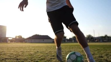 Profesyonel futbolcunun erkek ayağı, yeşil sahada koşarken topla numaralar gösterir. Sporcu bacakları gün batımında çayırda koşarken topa vurur. Stadyumda antrenman yapan genç adam. Yavaş çekim