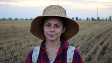 Mutlu gülümseyen dişi çiftçi buğday tarlasında kameraya bakıyor. Hasır şapkalı, arka planda arpa çayırı olan genç ve güzel tarımcı portresi. Tarım sektörü kavramı.