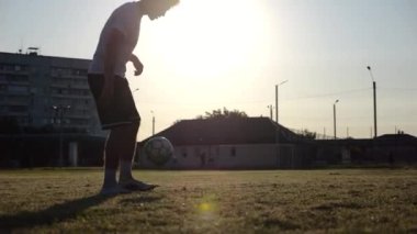 Günbatımında stadyumda futbol topu hokkabazlığı yapan profesyonel bir futbolcu. Genç adam yeşil sahada topa vuruyor. Sporcu arka planda güneş ışığıyla çayırda numaralar yapıyor. Serbest stil futbol kavramı.