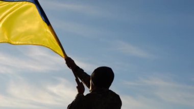 Arka planda mavi gökyüzüne karşı Ukrayna bayrağı sallayan üniformalı bir asker. Erkek Ukraynalı asker, Rus saldırısına karşı kazanılan zaferin şerefine ulusal bayrağı kaldırıyor..