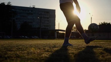 Yeşil sahada topa vuran genç adam bacakları. Günbatımında stadyumda futbol oynayan erkek futbolcu. Sporcu dışarıda numaralar yapıyor. Serbest stil futbol kavramı.
