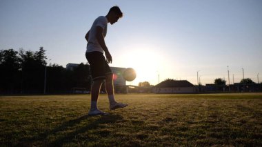 Günbatımında stadyumda futbol topu hokkabazlığı yapan profesyonel bir futbolcu. Genç adam yeşil sahada topa vuruyor. Sporcu arka planda güneş ışığıyla çayırda numaralar yapıyor. Serbest stil futbol kavramı.