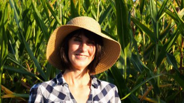 Mutlu gülümseyen dişi çiftçi mısır tarlasının yanında duran kameraya bakıyor. Hasır şapkalı, arka planda mısır tarlası olan yetişkin, güzel tarımcı portresi. Tarım sektörü kavramı.