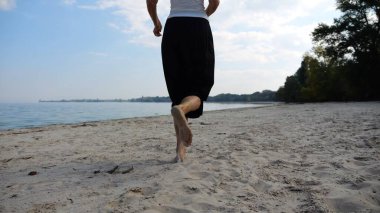 Sahilde arka planda deniz manzarasıyla koşan çıplak ayaklı bir kadın. Deniz kıyısında koşan dişi bacaklar. Mutlu bayan okyanus kıyısında vakit geçiriyor. Yaz tatili kavramı.