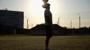 Günbatımında stadyumda futbol topu hokkabazlığı yapan profesyonel bir futbolcu. Genç adam yeşil sahada topa vuruyor. Sporcu arka planda güneş ışığıyla çayırda numaralar yapıyor. Serbest stil futbol kavramı.