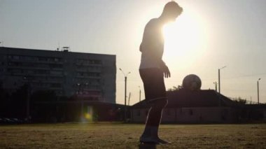 Günbatımında stadyumda futbol topu hokkabazlığı yapan profesyonel bir futbolcu. Genç adam yeşil sahada topa vuruyor. Sporcu arka planda güneş ışığıyla çayırda numaralar yapıyor. Serbest stil futbol kavramı.