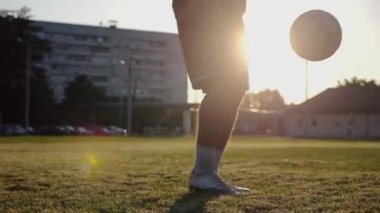 Günbatımında stadyumda futbol topu hokkabazlığı yapan profesyonel bir futbolcu. Genç adam yeşil sahada topa vuruyor. Sporcu arka planda güneş ışığıyla çayırda numaralar yapıyor. Serbest stil futbol kavramı.