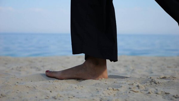 Female feet walking on sand at the beach with seascape at background. Legs of adult woman stepping along sea shore. Barefoot lady strolling near ocean coast. Concept of summer vacation