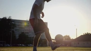 Günbatımında stadyumda futbol topu hokkabazlığı yapan profesyonel bir futbolcu. Genç adam yeşil sahada topa vuruyor. Sporcu arka planda güneş ışığıyla çayırda numaralar yapıyor. Serbest stil futbol kavramı.