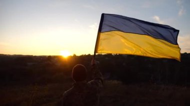 Askeri üniformalı genç adam arka planda gün batımına karşı Ukrayna bayrağı sallıyor. Erkek Ukraynalı asker kırsal kesimde ulusal bayrağı kaldırdı. Rus saldırısına karşı zafer. Savaşın sonu..