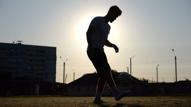 Günbatımında stadyumda futbol topu hokkabazlığı yapan profesyonel bir futbolcu. Genç adam yeşil sahada topa vuruyor. Sporcu arka planda güneş ışığıyla çayırda numaralar yapıyor. Serbest stil futbol kavramı.