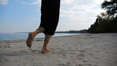 Sahilde arka planda deniz manzarasıyla koşan çıplak ayaklı bir kadın. Deniz kıyısında koşan dişi bacaklar. Mutlu bayan okyanus kıyısında vakit geçiriyor. Yaz tatili kavramı.