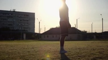 Günbatımında stadyumda futbol topu hokkabazlığı yapan profesyonel bir futbolcu. Genç adam yeşil sahada topa vuruyor. Sporcu arka planda güneş ışığıyla çayırda numaralar yapıyor. Serbest stil futbol kavramı.