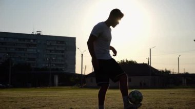 Günbatımında stadyumda futbol topu hokkabazlığı yapan profesyonel bir futbolcu. Genç adam yeşil sahada topa vuruyor. Sporcu arka planda güneş ışığıyla çayırda numaralar yapıyor. Serbest stil futbol kavramı.