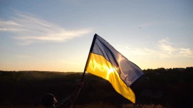 Erkek Ukrayna askeri, arkasında günbatımı olan bir Ukrayna bayrağı kaldırdı. Kamuflaj üniformalı genç adam, Rus saldırısına karşı kazanılan zaferin şerefine mavi-sarı bayrak kaldırdı..