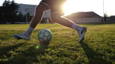 Profesyonel futbolcunun erkek ayağı, yeşil sahada koşarken topla numaralar gösterir. Sporcu bacakları gün batımında çayırda koşarken topa vurur. Stadyumda antrenman yapan genç adam. Yavaş çekim