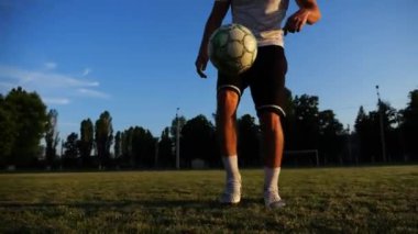 Günbatımında stadyumda futbol topu hokkabazlığı yapan profesyonel bir futbolcu. Genç adam yeşil sahada topa vuruyor. Sporcu arka planda güneş ışığıyla çayırda numaralar yapıyor. Serbest stil futbol kavramı.