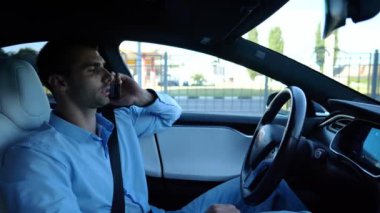 Şehir yolunda otomatik pilotla elektrikli otomobil sürerken akıllı telefondan konuşan erkek girişimci. Kendine güvenen işadamı, otomatik elektrikli araba kullanırken telefonla konuşuyor..