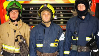 Büyük kırmızı bir arabanın yanında duran kasklı ve koruyucu üniformalı erkek itfaiyecilerin portresi. Tam teçhizatlı genç itfaiyeciler istasyondaki itfaiye arabasının arka planında poz veriyorlar..