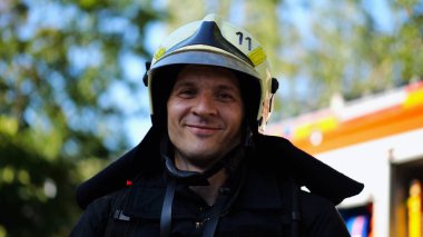 Portrait of young confident fireguard in protective uniform against background of a fire truck. Male firefighter in full equipment looking into camera with positive emotions near big red car. clipart