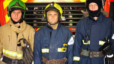 Portrait of male firefighters in helmets and protective uniforms standing near a big red car. Young confident firemen in full equipment posing against the background of a fire engine at station. clipart