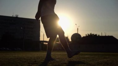 Günbatımında stadyumda futbol topu hokkabazlığı yapan profesyonel bir futbolcu. Genç adam yeşil sahada topa vuruyor. Sporcu arka planda güneş ışığıyla çayırda numaralar yapıyor. Serbest stil futbol kavramı.