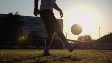 Yeşil sahada topa vuran genç adam bacakları. Günbatımında stadyumda futbol oynayan erkek futbolcu. Sporcu dışarıda numaralar yapıyor. Serbest stil futbol kavramı.