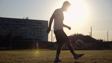Günbatımında stadyumda futbol topu hokkabazlığı yapan profesyonel bir futbolcu. Genç adam yeşil sahada topa vuruyor. Sporcu arka planda güneş ışığıyla çayırda numaralar yapıyor. Serbest stil futbol kavramı.