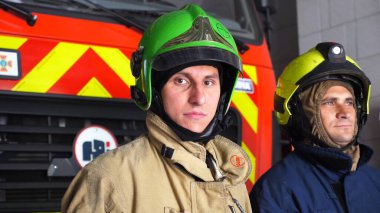 Bölümdeki büyük kırmızı arabanın yanında duran kasklı ve koruyucu üniformalı erkek itfaiyecilerin portresi. Tam teçhizatlı genç itfaiyeciler istasyondaki itfaiye arabasının arka planında poz veriyorlar..