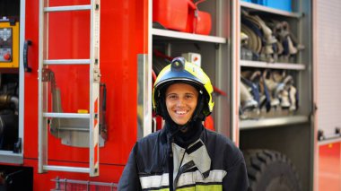 Portrait of happy female fireguard against the background of big red truck. Young firewoman in full equipment looking at camera with positive emotions near a fire engine. Concept of heroic profession. clipart