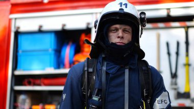 Portrait of confident male fireguard with dirty face standing near a fire engine. Young fireman in full equipment posing after fire against the background of big red truck. Concept of saving lives. clipart