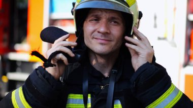 Young fireguard in protective uniform putting helmet against background of a big red truck. Male firefighter in full equipment near fire engine ready to work. Saving lives and heroic profession. clipart