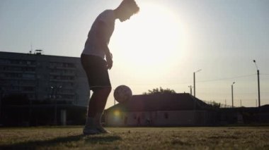 Günbatımında stadyumda futbol topu hokkabazlığı yapan profesyonel bir futbolcu. Genç adam yeşil sahada topa vuruyor. Sporcu arka planda güneş ışığıyla çayırda numaralar yapıyor. Serbest stil futbol kavramı.