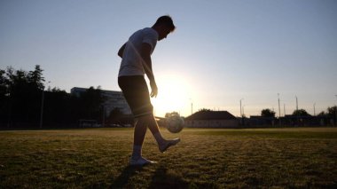Günbatımında stadyumda futbol topu hokkabazlığı yapan profesyonel bir futbolcu. Genç adam yeşil sahada topa vuruyor. Sporcu arka planda güneş ışığıyla çayırda numaralar yapıyor. Serbest stil futbol kavramı.
