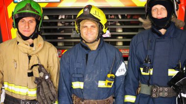 Korumalı üniformalı genç itfaiyecilerin portresi istasyondaki itfaiye aracının arka planında. Tam teçhizatlı erkek itfaiyeciler büyük kırmızı arabanın yanında pozitif duygularla kameraya bakıyorlar..