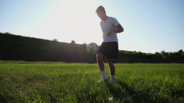 Güneşli bir günde stadyumda futbol topuyla hokkabazlık yapan sporcu. Genç adam yeşil sahada topa vuruyor. Profesyonel futbolcu, arka planda güneş ışığıyla çayırda numaralar yapıyor. Serbest stil futbol.