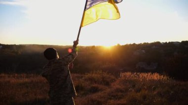 Ukraynalı asker gün batımının arka planına ulusal bayrak sallıyor. Genç erkek asker kırsal kesimde bayrak kaldırdı. Rus saldırısına karşı zafer. Rusya ve Ukrayna arasındaki savaşın sonu..
