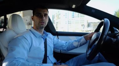 Şehir yolunda modern elektrikli araba kullanan yakışıklı bir işadamının profili. Erkek başarılı girişimci şehirde elektrikli araçla geziyor. Genç adam güneşli bir günde işe gidip geliyor. Yavaş çekim