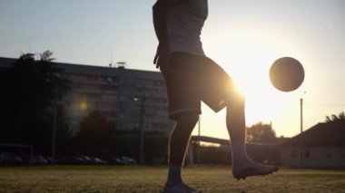 Günbatımında stadyumda futbol topu hokkabazlığı yapan profesyonel bir futbolcu. Genç adam yeşil sahada topa vuruyor. Sporcu arka planda güneş ışığıyla çayırda numaralar yapıyor. Serbest stil futbol kavramı.