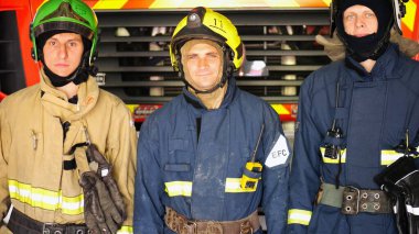 Büyük kırmızı bir arabanın yanında duran kasklı ve koruyucu üniformalı erkek itfaiyecilerin portresi. Tam teçhizatlı genç itfaiyeciler istasyondaki itfaiye arabasının arka planında poz veriyorlar..