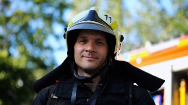 Portrait of young confident fireguard in protective uniform against background of a fire truck. Male firefighter in full equipment looking into camera with positive emotions near big red car. clipart